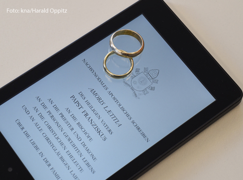 Auf einem Tablet ist das päpstliche Lehrschreiben "Amoris laetitia" geöffnet. Darüber liegen zwei Eheringe. 