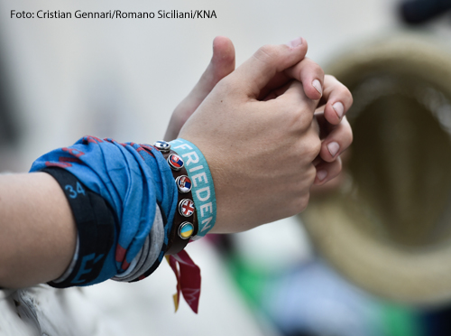	Betende Hände eines Jugendlichen am 31. Juli 2018 im Vatikan. Am Handgelenk trägt er ein Band mit der Aufschrift "Frieden".