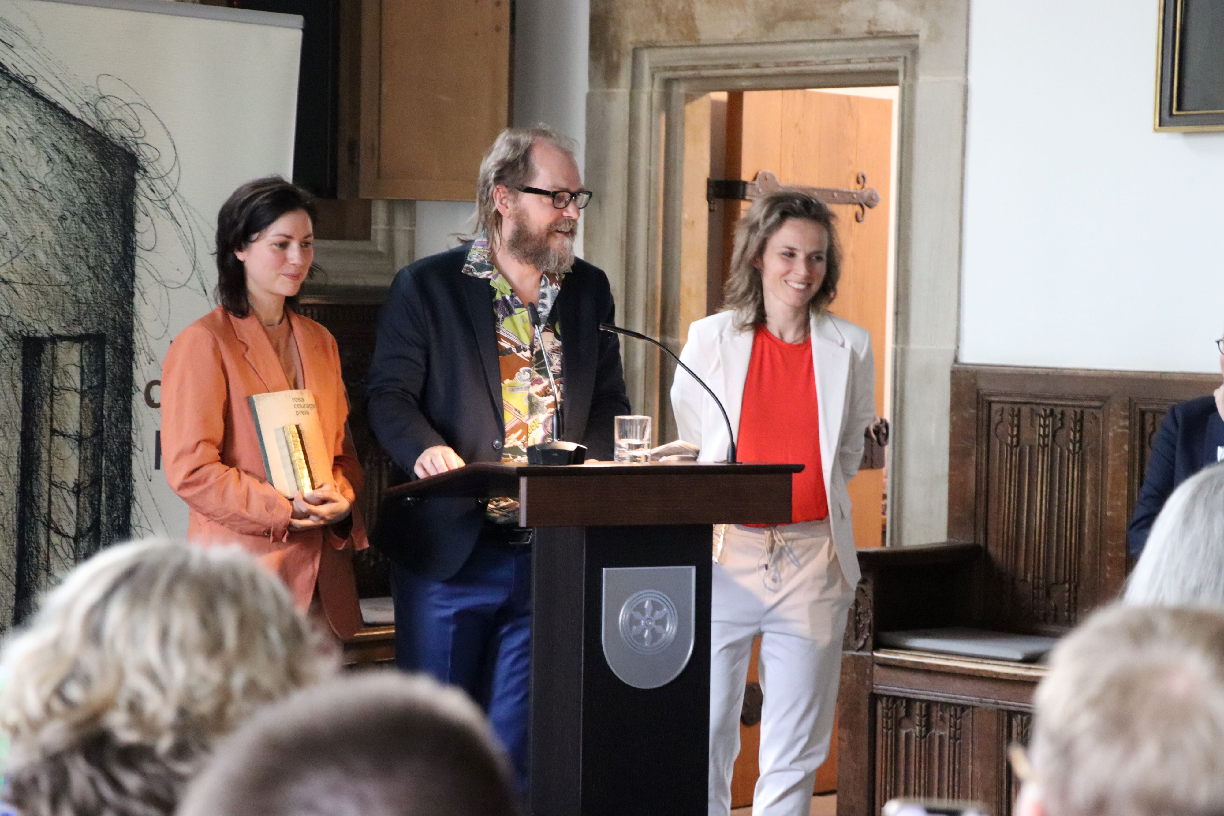 Ein Mann steht zwischen zwei Frauen am Rednerpult. Eine Frau hält einen Preis in der Hand