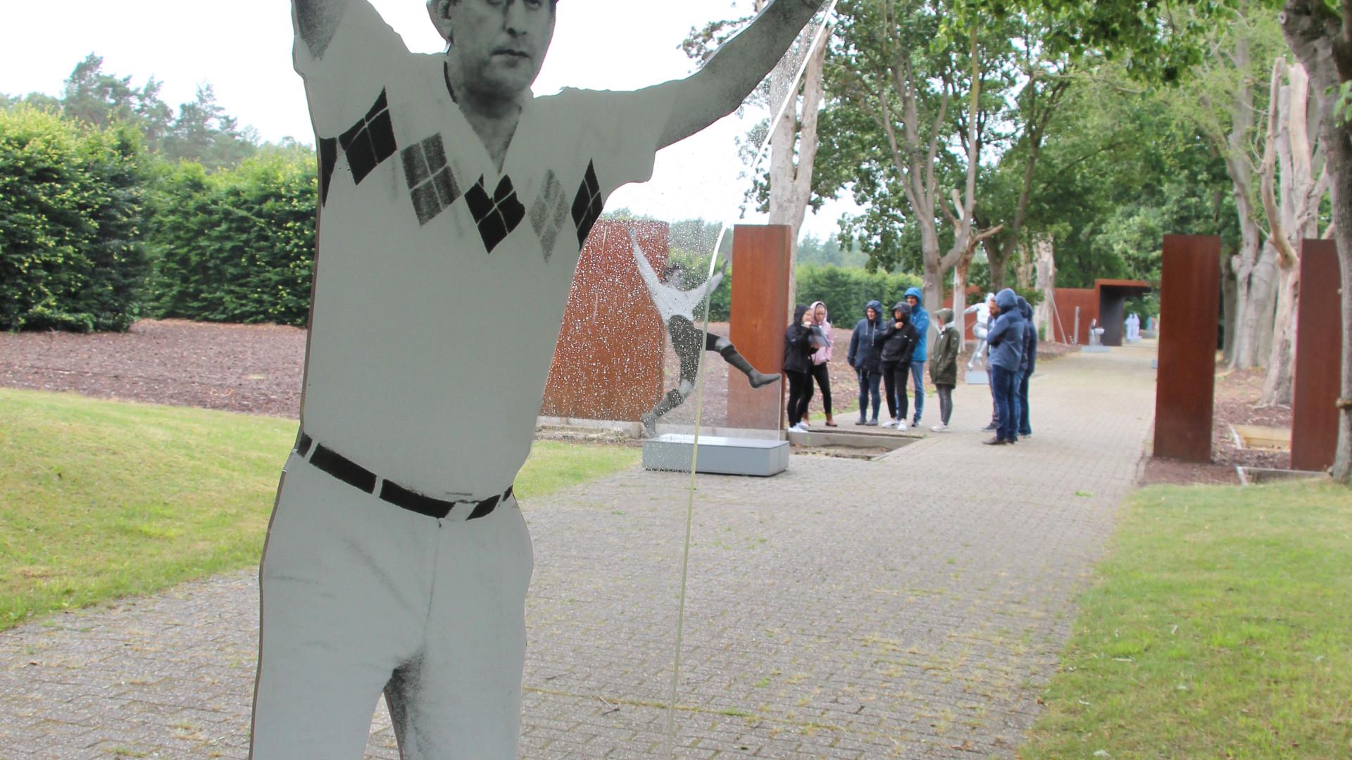 Eine Figur steht auf einem Weg und hebt die Hände. Im Hintergrund ist eine Gruppe zu sehen. 