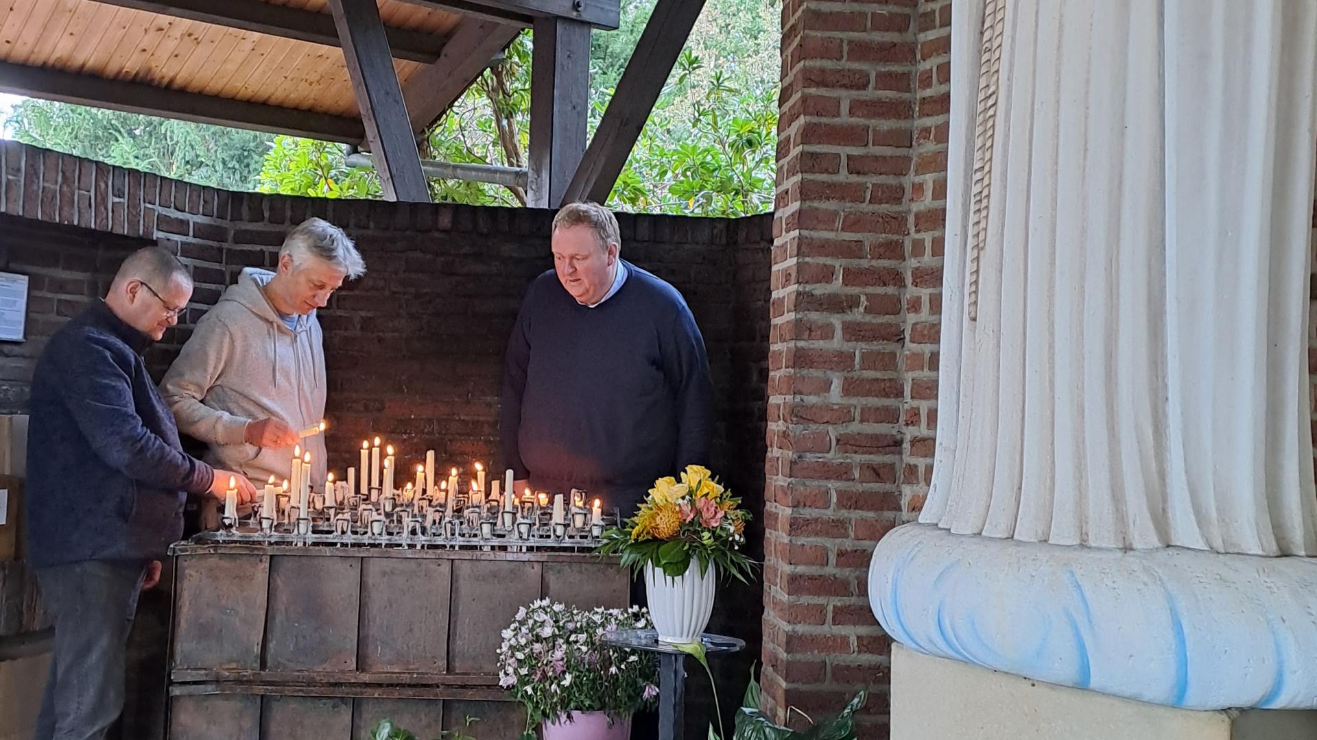 Blumen stehen vor der Muttergottes-Statue. Im Hintergrund zünden drei Männer Kerzen an.