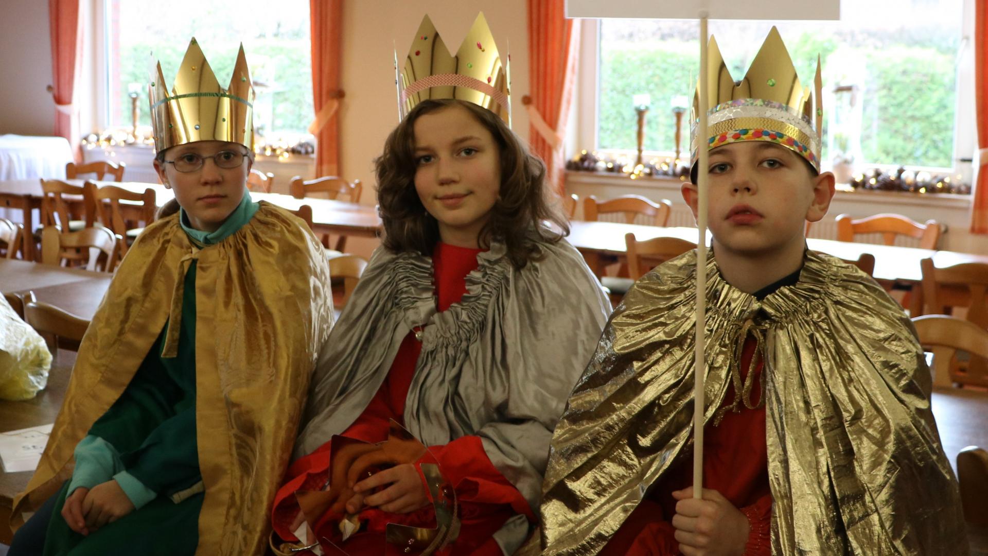 Drei Kinder sitzen in bunten Umhängen und mit Kronen auf dem Kopf auf einem Tisch. 