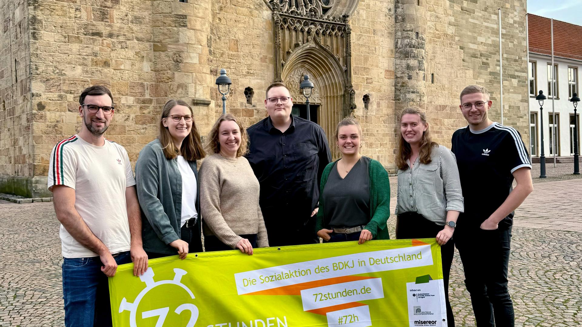 Sienen junhge Menschen haben sich hinter einem Transpaent aufgestellt, das auf die 72-Stunden-Aktion hinweist. Im Hintergrund ist der Dom zu sehen.
