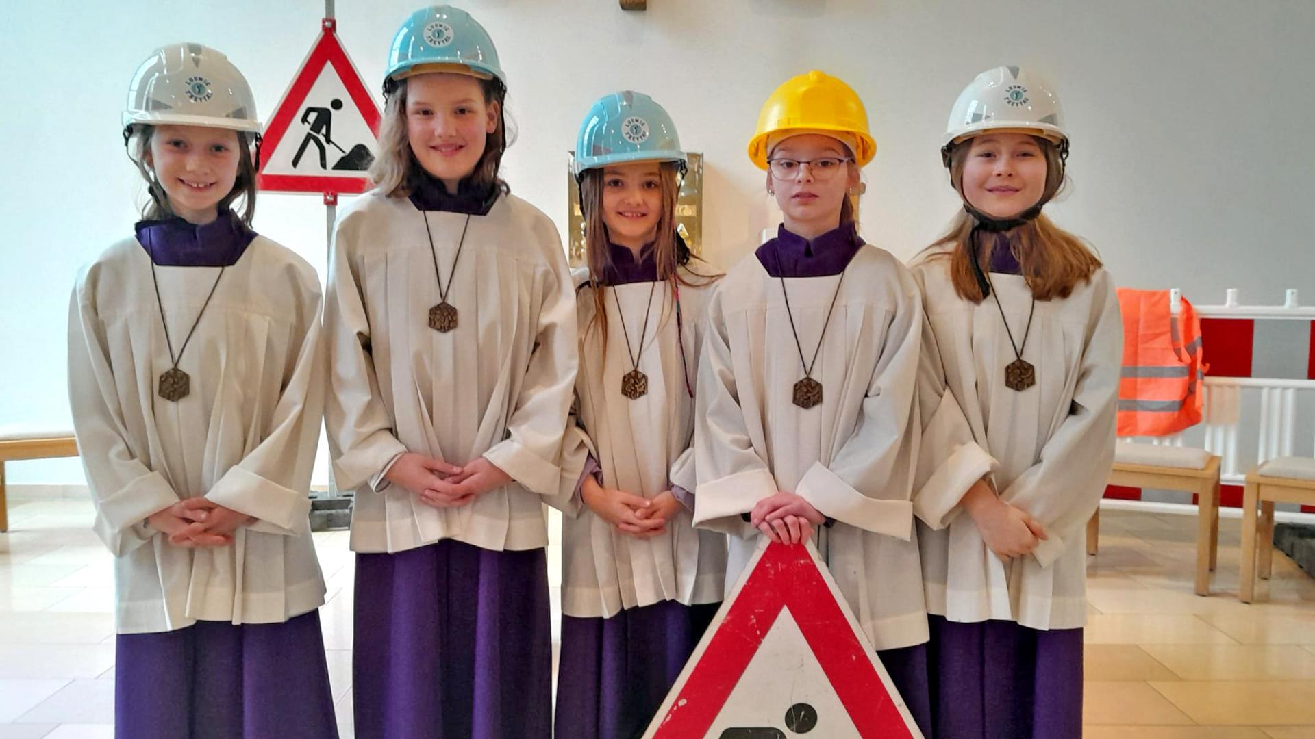 Fünf Kinder stehen in Messdienergewändern und Bauhelmen auf dem Kopf vor einem Baustellenschild