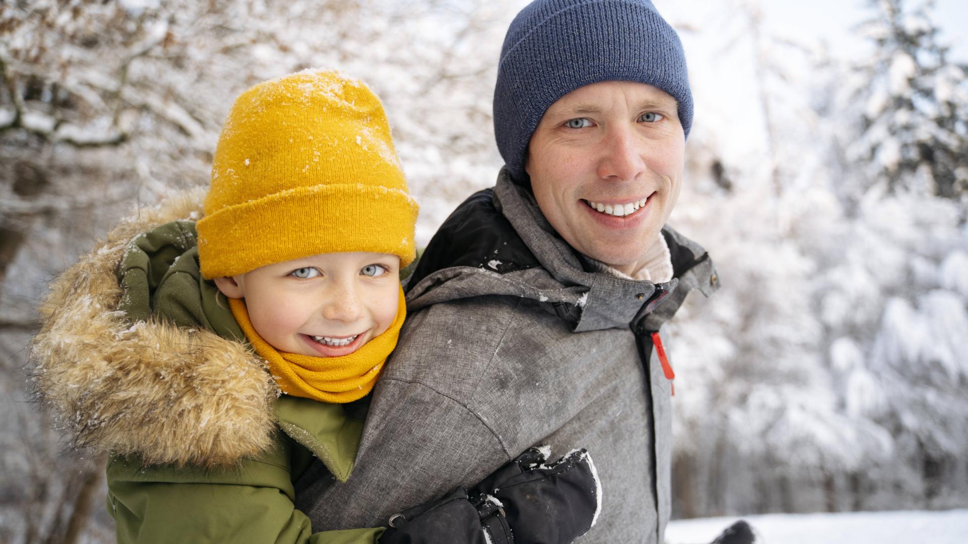 Ein Vater trägt sein Kindergartenkind huckepack auf dem Rücken, beide schauen fröhlich in die Kamera.