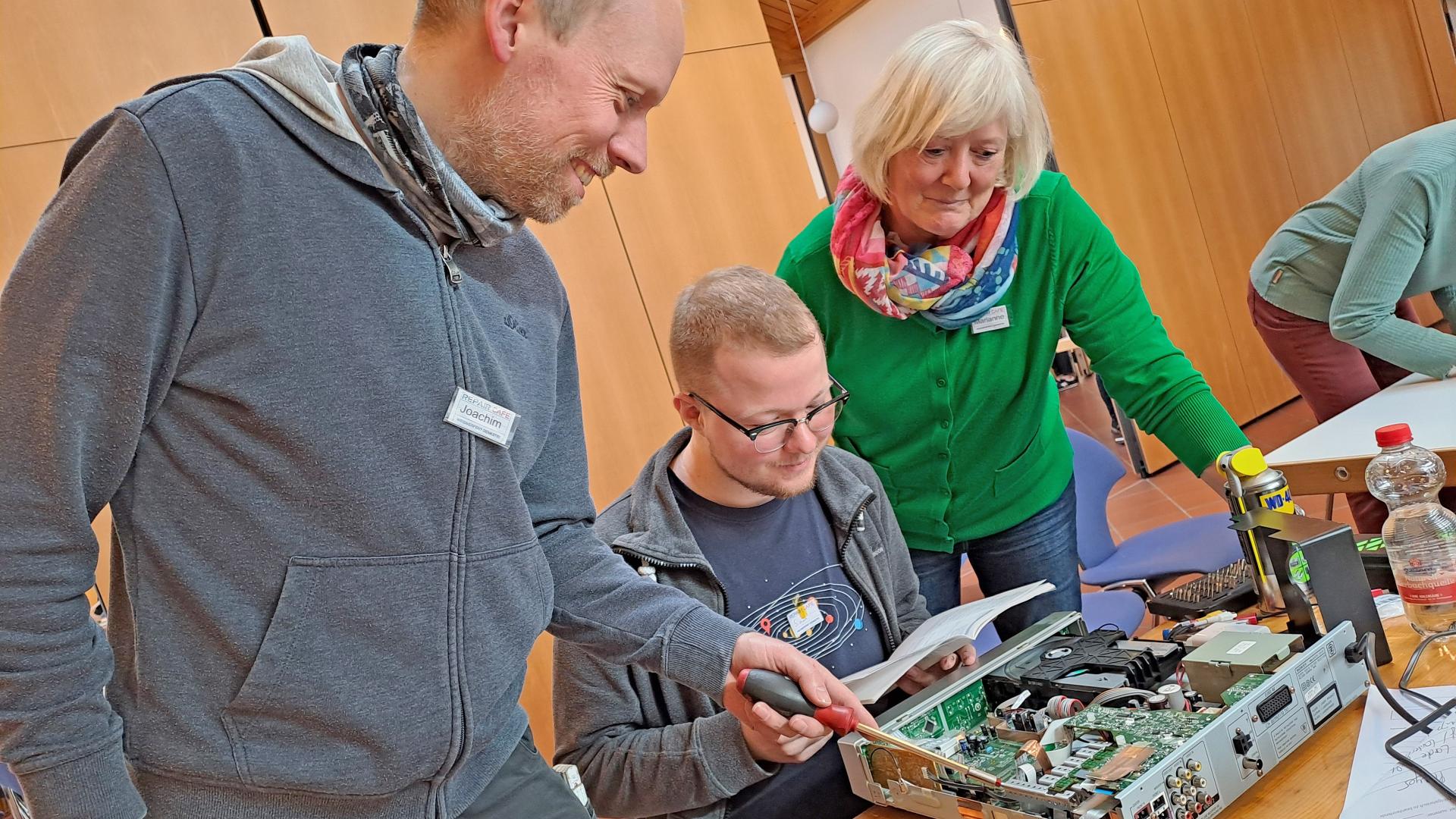 Ein junger Mann sitzt vor einem aufgeschraubten elektronischen Gerät. Zwei weitere Personen stehen daneben und nutzen einen Schraubenzieher