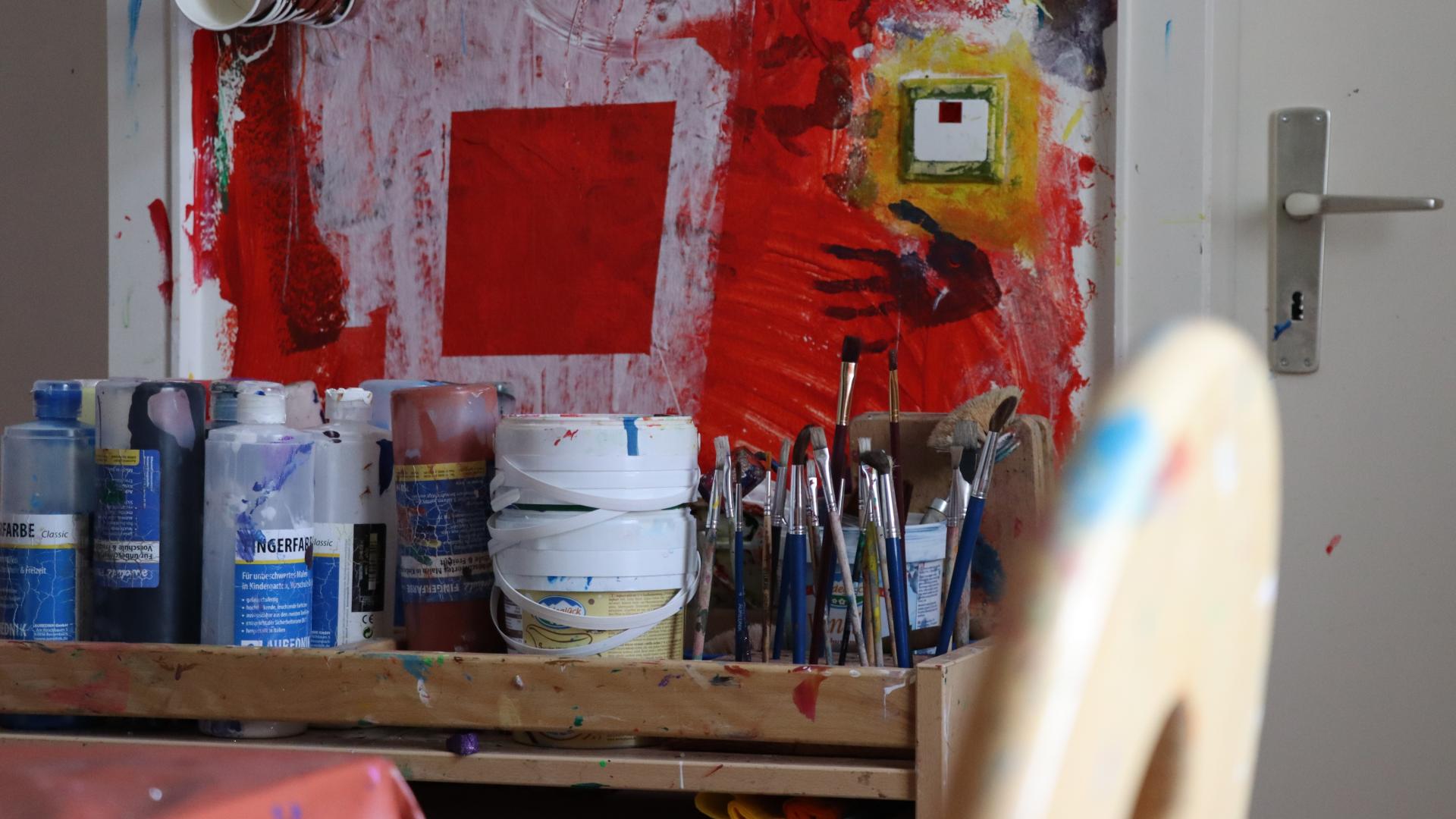 Vor einer Bunt bemalten Wand stehen viele kleine Töpfe und Pinsel