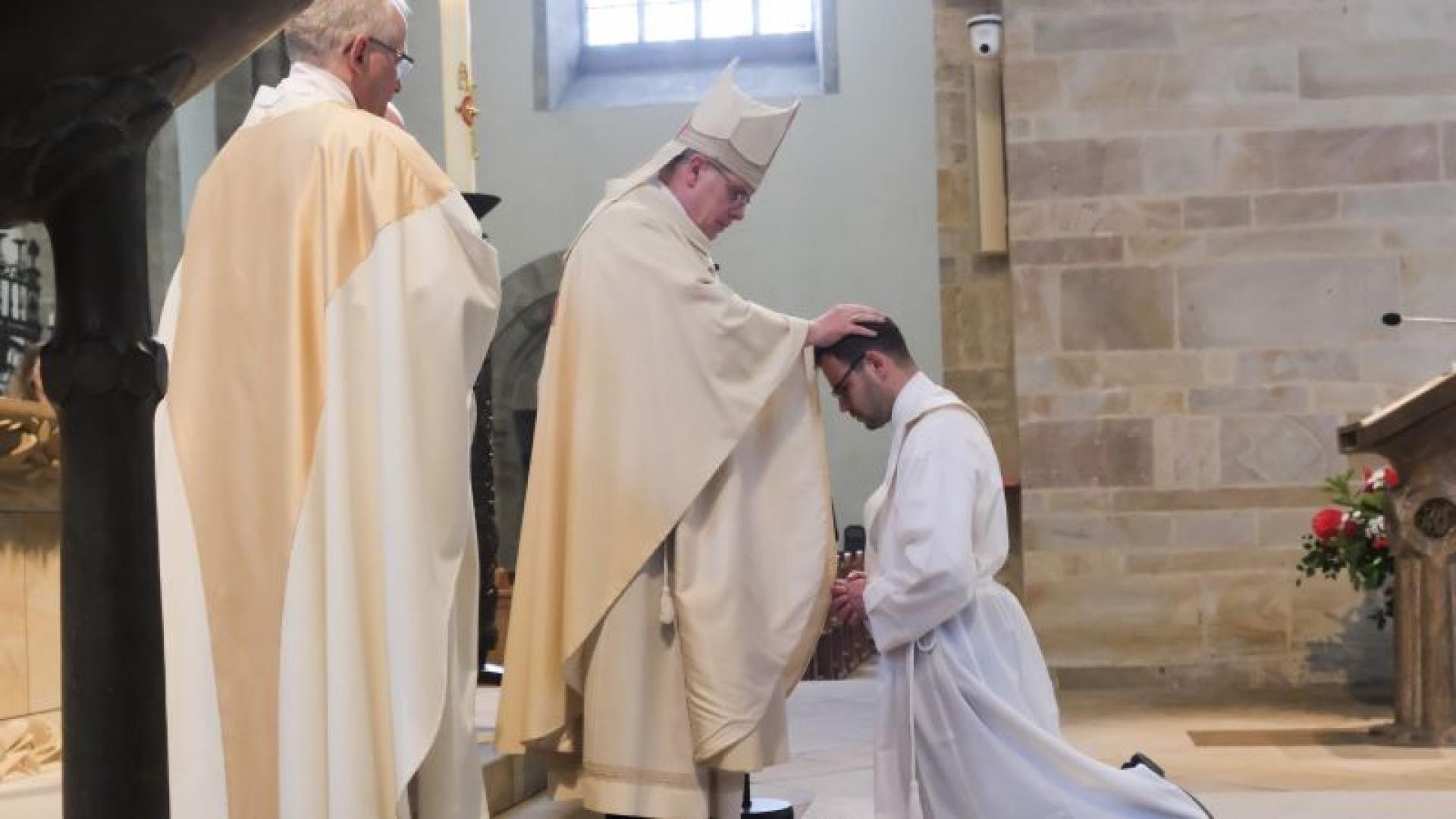 Ein Bischof legt einem vor ihm knieenden Diakon die Hände auf, um ihn zum Priester zu weihen.